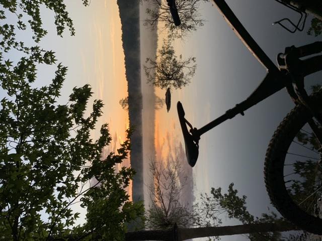A scenic view at dawn featuring a calm body of water reflecting a colorful sunrise. In the foreground, the silhouette of a bicycle is visible, framed by green leaves and trees, while mist rises gently from the water's surface. Allatoona Creek Park mountain bike trail.