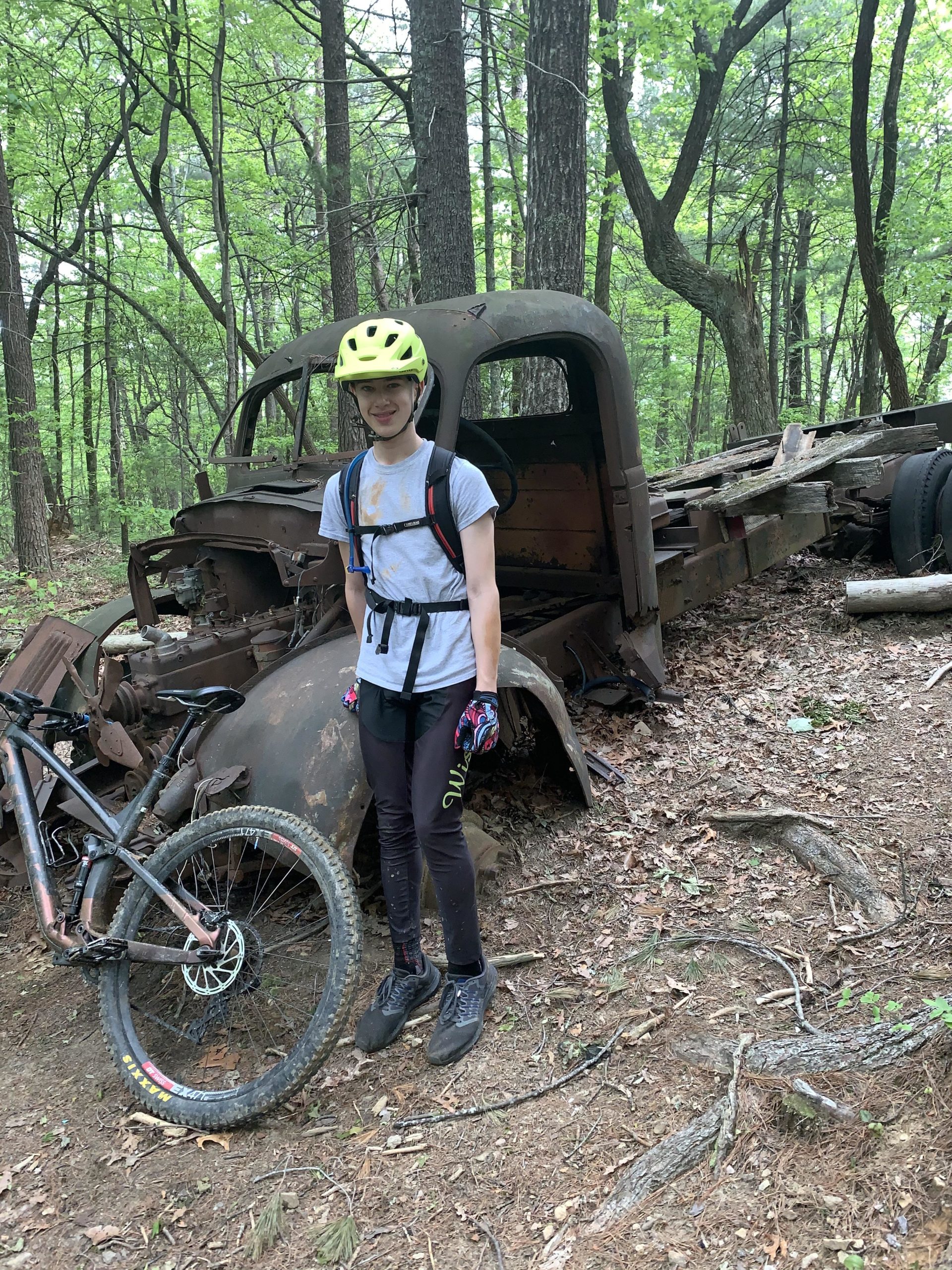 A young person wearing a bright yellow helmet and gloves stands beside a partially rusted, abandoned truck in a forested area. A mountain bike is leaned against the truck, and the ground is covered with leaves and twigs. The surroundings feature tall trees with fresh green foliage, suggesting a lush, natural environment. Bull / Jake Mountain mountain bike trail.