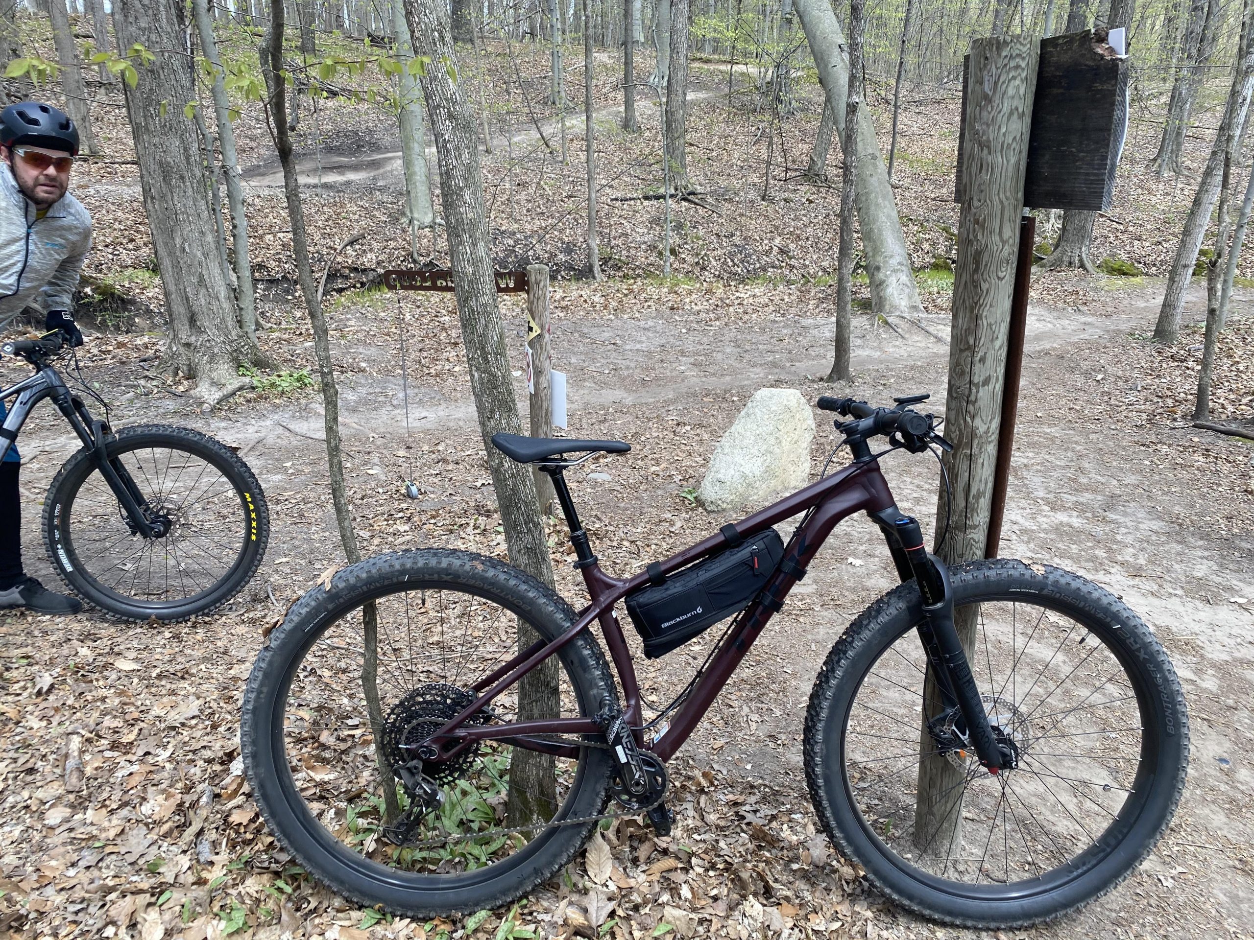 Trek Stache 7: A mountain bike with a maroon frame is parked next to a wooden signpost in a forested area, surrounded by autumn leaves. A cyclist in a gray jacket and helmet is seen beside another black mountain bike in the background. The scene depicts a trail with trees and a rocky path visible in the distance.