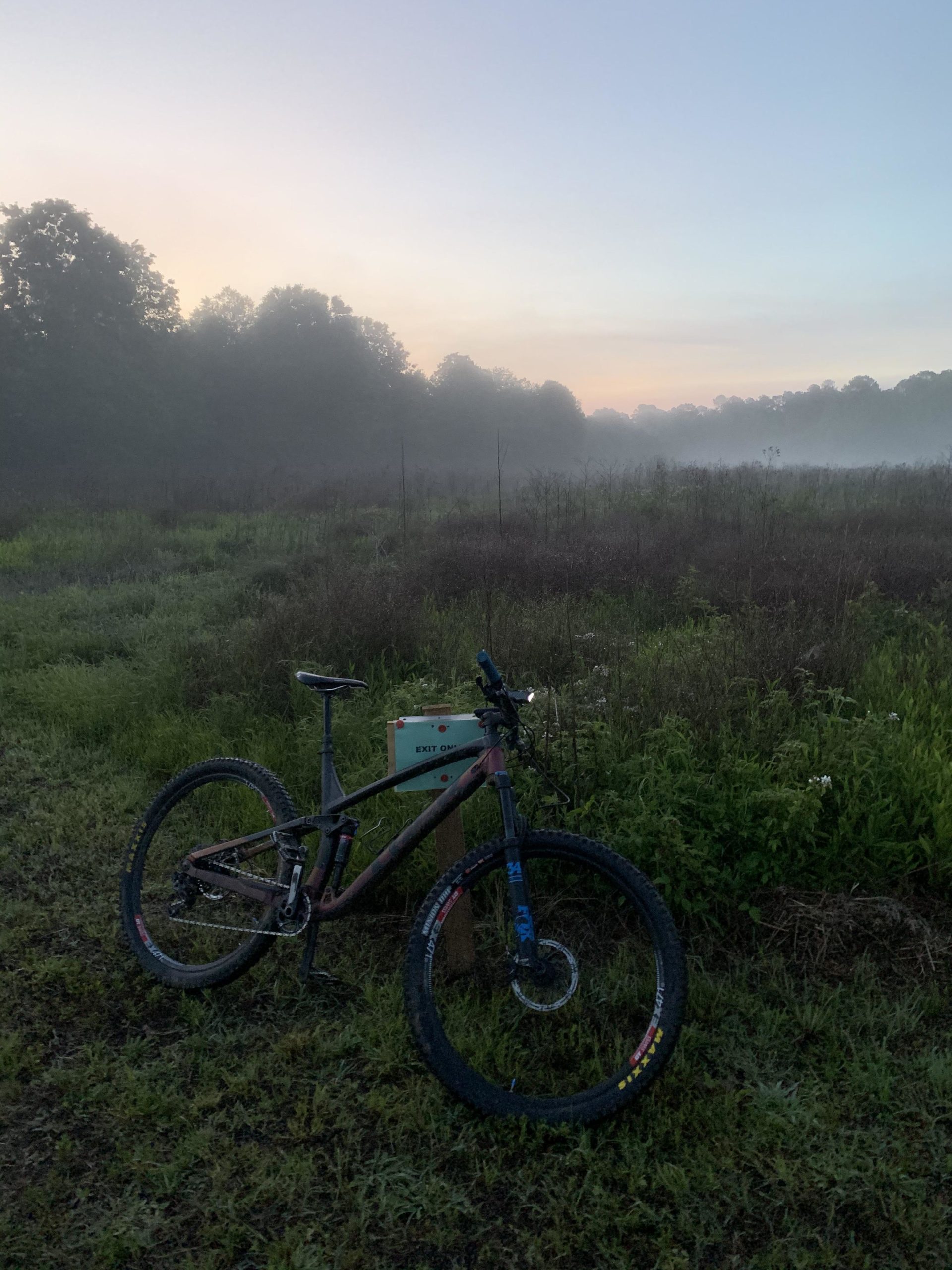 A mountain bike resting on the grass in a misty field at dawn, with trees and a soft, colorful sky in the background. A small sign labeled "EXIT ONE" is visible nearby. The scene conveys a peaceful and serene atmosphere. Allatoona Creek Park mountain bike trail.