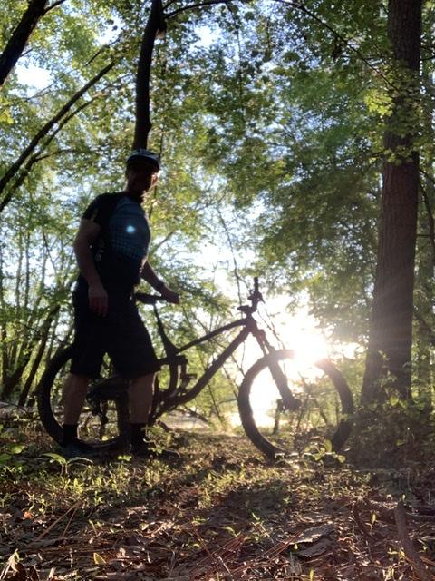 A silhouetted figure stands next to a mountain bike in a wooded area, with sunlight filtering through the trees in the background. The scene captures a tranquil outdoor moment, surrounded by greenery and soft light. Allatoona Creek Park mountain bike trail.
