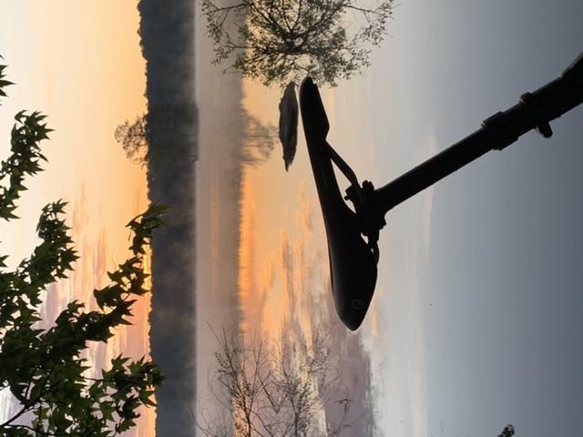 A tranquil sunrise reflected on a calm body of water, with mist rising above the surface. In the foreground, a bicycle seat and handlebars silhouette against the vibrant colors of the sky, framed by leaves from nearby trees. Allatoona Creek Park mountain bike trail.