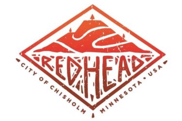 Logo of the Redhead organization, featuring a stylized mountain design and the text "REDHEAD" prominently displayed in bold letters. The logo is framed within a diamond shape and includes the words "CITY OF CHISHOLM, MINNESOTA, USA" at the bottom. The color scheme consists of warm shades of red and orange. Redhead MTB Park mountain bike trail.