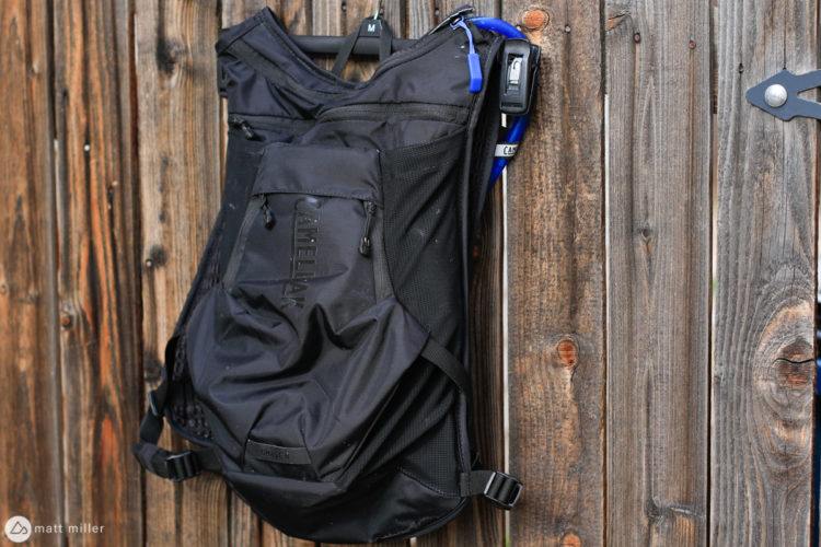 A black CamelBak backpack hanging on a wooden fence, featuring mesh side pockets and a hydration tube.