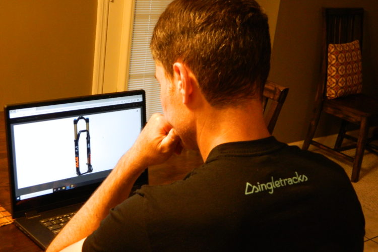 Person sitting at a desk, viewing a bicycle fork image on a laptop screen. The individual has short hair and is wearing a black shirt with "singletracks" printed on the back. A chair with a patterned cushion is visible in the background. The setting is cozy and well-lit with natural light coming from a window.
