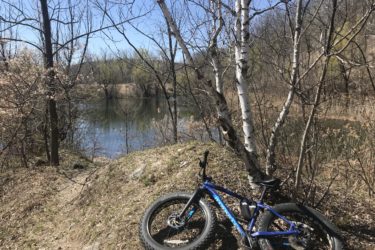 A blue fat bike resting against a birch tree near a calm body of water, surrounded by trees and budding vegetation under a clear blue sky. Glacial Blue Hills mountain bike trail.