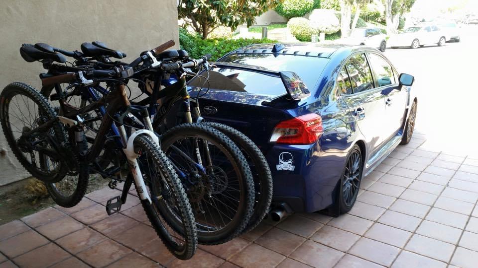 Specialized Stumpjumper FSR Expert 29: A blue car parked on a tiled driveway with three mountain bikes mounted on a rear bike rack. The setting includes greenery and other parked cars in the background.