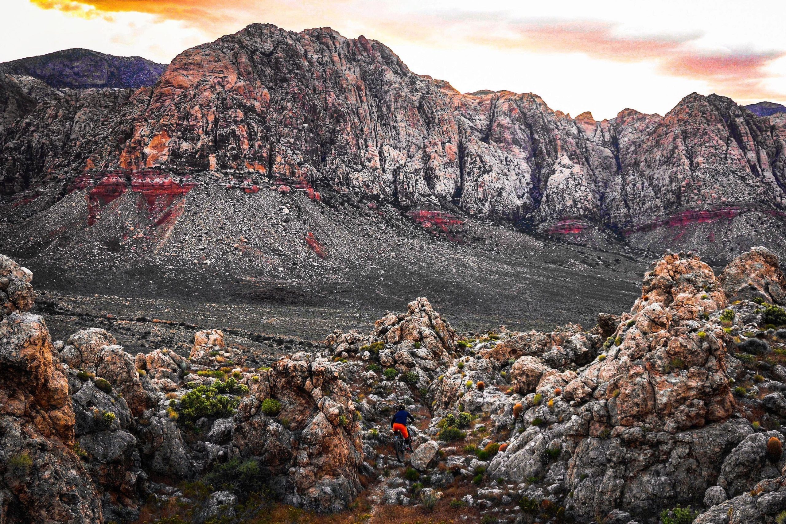 A cyclist riding through rocky terrain with colorful mountainous backdrop during sunset. The landscape features rugged rocks and scattered vegetation, displaying a mix of earthy tones against the vibrant sky. Cottonwood Valley North mountain bike trail.