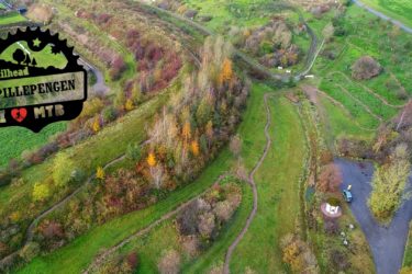 Aerial view of a mountain biking trailhead at Spillepengen, featuring winding paths through vibrant greenery and trees. The scene includes a parking area and a trail map stand, with the surrounding landscape showcasing autumn colors. A logo in the corner highlights the trailhead's identity, emphasizing its appeal for mountain biking enthusiasts. Red Trail mountain bike trail.