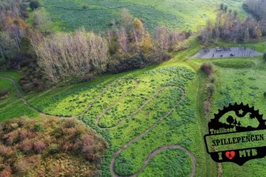 Aerial view of an outdoor mountain biking trail, featuring winding paths cut through lush green grass and surrounded by trees. A small picnic area with tables is visible nearby, along with a logo that reads "Trailhead Spillepeng" and "I ❤️ MTB" in the corner. Red Trail mountain bike trail.