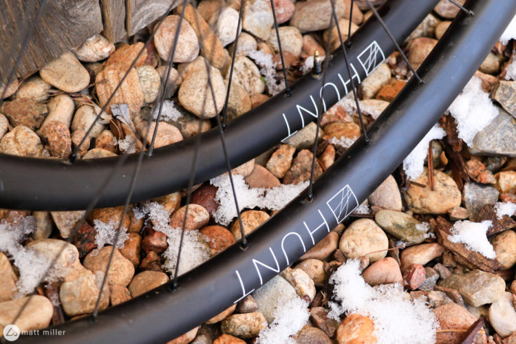 Two black bicycle wheels partially resting on a bed of small, round stones, with a light dusting of snow scattered among the rocks. The wheels feature the brand name "HUNT" displayed in white lettering.