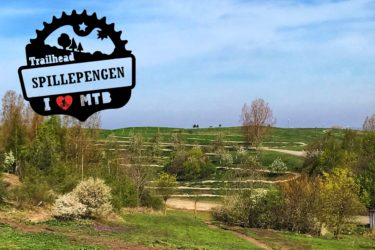 A scenic view of a mountain bike trailhead at Spillepengen, featuring rolling green hills, trees, and a clear blue sky. The image includes a graphic overlay that reads "Trailhead Spillepengen I ❤️ MTB," indicating a mountain biking location. Red Trail mountain bike trail.