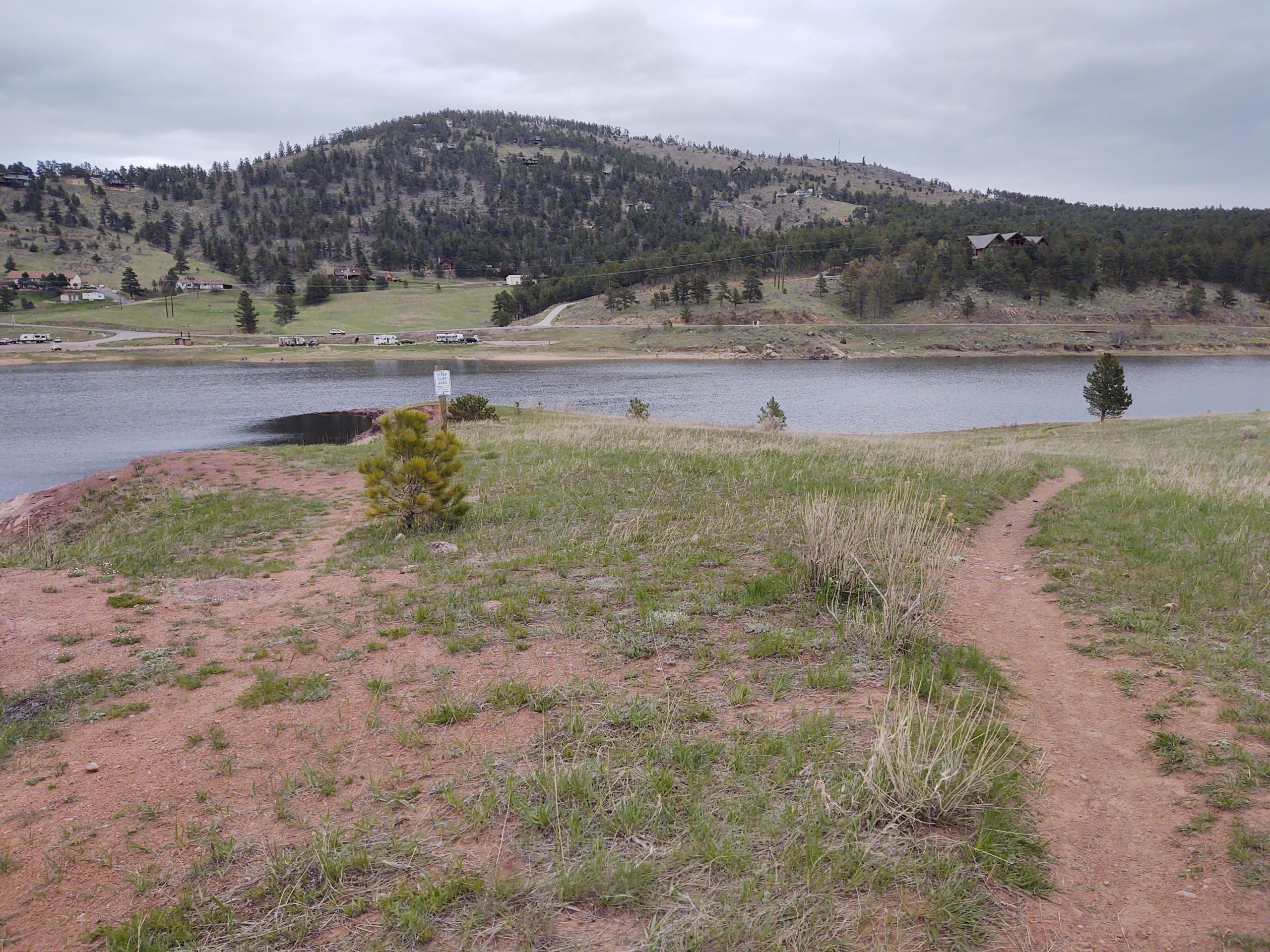 A scenic landscape featuring a winding dirt path leading towards a tranquil lake, surrounded by green grass and patches of light brown earth. The backdrop includes rolling hills dotted with trees, and a cloudy sky overhead, creating a serene, natural setting. In the distance, a few houses and vehicles are visible along the shoreline. Pinewood Reservoir mountain bike trail.