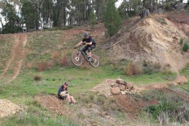 A mountain biker performing a jump on a dirt track while a second person sits nearby on a mound of dirt, watching the action in a natural outdoor setting. Surrounding the scene are trees and earthy terrain. Geurie Goat Track mountain bike trail.