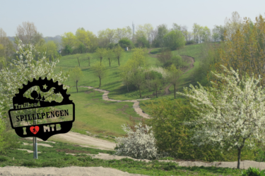 A scenic view of a green hillside with winding trails, showcasing a trailhead sign that reads "Trailhead Spillepengen I ❤️ MTB." The landscape features blooming trees and well-maintained paths ideal for mountain biking. Blue Trail mountain bike trail.