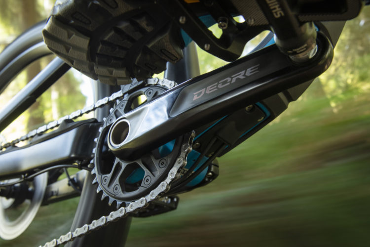 Close-up view of a mountain bike's crankset and chain, showcasing a Shimano Deore component, with a blurred background indicating motion through a natural landscape.
