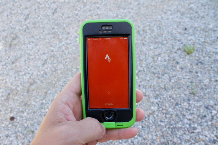 A hand holding a smartphone with a bright red screen displaying the logo of an app, set against a background of gravel. The phone is encased in a green protective case.