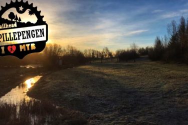 A serene landscape at sunset featuring a grassy area with trees in the distance. A gentle stream reflects the warm colors of the sky. The image includes a logo in the corner that reads "Trailhead Spillepengen" with the phrase "I ❤️ MTB." Blue Trail mountain bike trail.