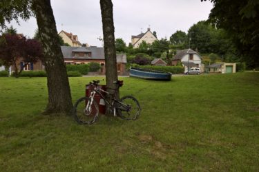 A scenic view of a grassy area with a bicycle leaning against a tree. In the background, there are several houses and a parked boat, with greenery and trees surrounding the area, under a cloudy sky. Hidenbourg line mountain bike trail.