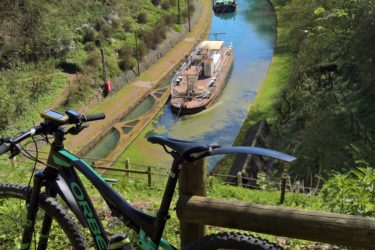 A mountain bike stands on a grassy hillside near a wooden fence, overlooking a serene canal surrounded by lush green trees. Several boats are moored along the waterway, which reflects the clear blue sky above. The scene captures a peaceful outdoor setting, ideal for cycling and enjoying nature. Hidenbourg line mountain bike trail.