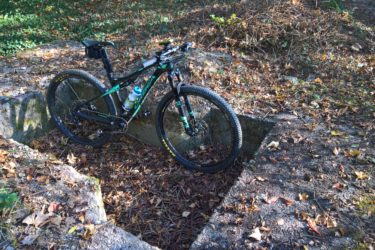A mountain bike resting on a stone foundation surrounded by fallen leaves and greenery in a natural outdoor setting. A water bottle is attached to the bike frame. Hidenbourg line mountain bike trail.