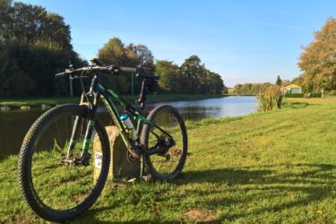 A mountain bike rests on a stone marker beside a calm river, surrounded by lush green grass and trees. The sky is clear and sunny, creating a peaceful outdoor scenery perfect for cycling or relaxing by the water. Hidenbourg line mountain bike trail.