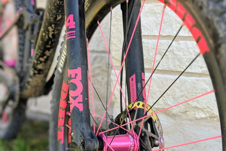 Close-up of a mountain bike's front wheel, featuring a black fork with pink accents and a dirty frame. The wheel has bright pink spokes and a yellow brake rotor, set against a textured white stone background.