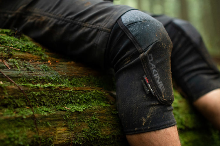 A close-up view of a knee pad resting against a moss-covered log in a forest setting, showcasing dirt and wear. The knee pad features the brand name "Dakine" prominently displayed.