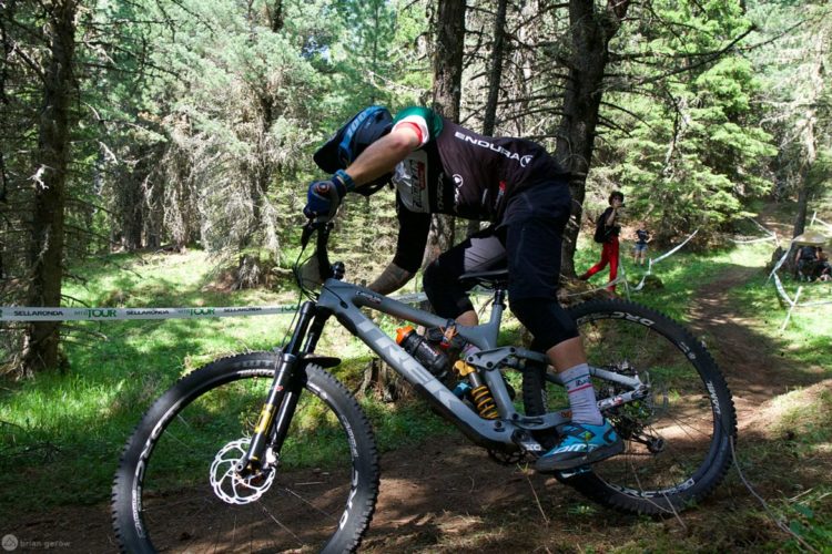 A mountain biker in a competitive stance navigating a wooded trail, surrounded by trees. The cyclist wears a helmet and protective gear, with a focus on balance and control as they maneuver over the terrain. In the background, spectators can be seen watching the event.