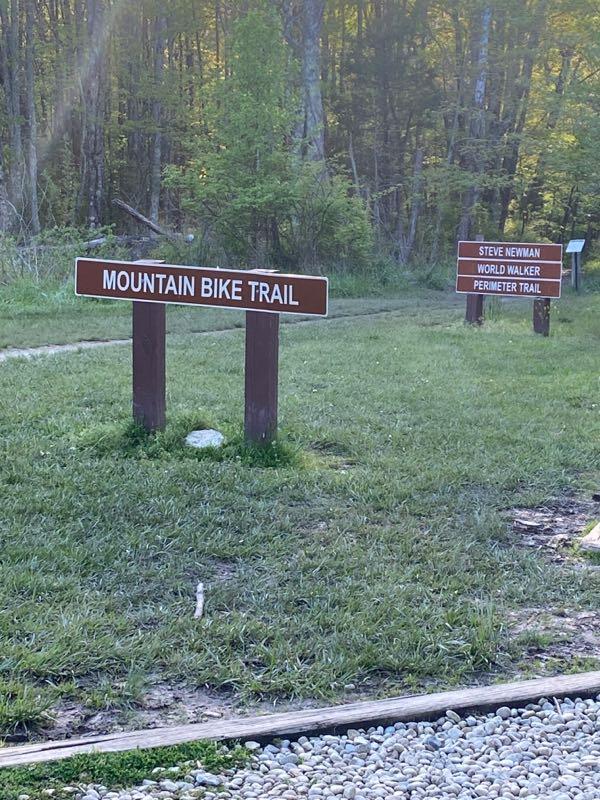 Two wooden signs in a grassy area indicating trail paths. The left sign reads "Mountain Bike Trail," while the right sign lists "Steve Newman World Walker Perimeter Trail." The background features trees with early morning light filtering through. East Fork mountain bike trail.