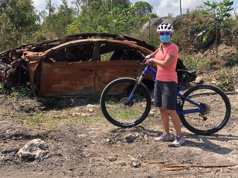 A person wearing a helmet and a face mask stands next to a blue mountain bike in front of a rusty, abandoned car in a natural setting with trees and shrubs in the background. Bird Basin Park mountain bike trail.