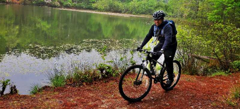 A person riding a mountain bike along a trail beside a calm lake, surrounded by lush greenery and trees. The scene captures a tranquil nature setting with reflections in the water. Tribble Mill Park mountain bike trail.