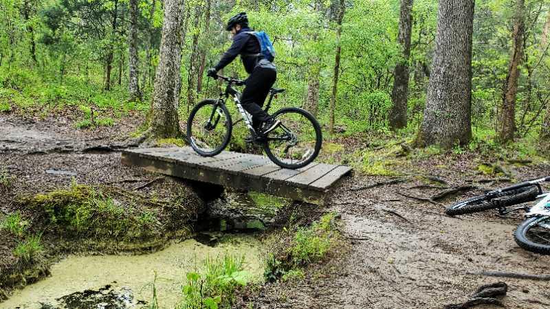 A person riding a mountain bike crosses a wooden bridge over a small stream in a forested area. The ground is muddy, and greenery surrounds the trail, creating a vibrant outdoor scene. Another bicycle is lying on the ground beside the path. Tribble Mill Park mountain bike trail.