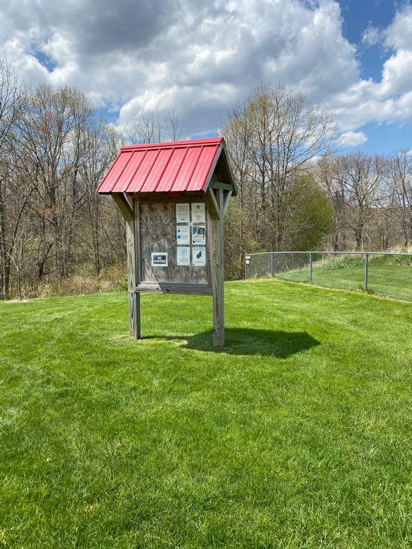 Wooden bulletin board with a red roof, displaying various signs and information, set on a grassy area surrounded by trees and a chain-link fence under a partly cloudy sky. Holly High School mountain bike trail.