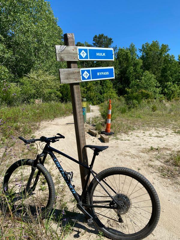 A mountain bike rests on the ground next to a wooden signpost indicating two trail options: "HULK" and "BYPASS." The background features a clear blue sky and lush greenery, with a traffic cone visible nearby, suggesting trail maintenance. The scene captures an outdoor setting ideal for biking or hiking. Horry County Bike Run Park mountain bike trail.