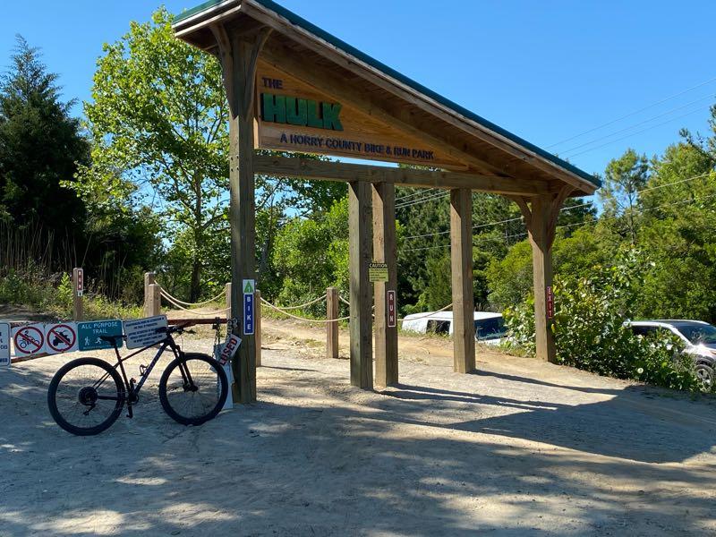 An entrance sign for "The Hulk," a biking and fun park in Horry County. The wooden structure features the park's name prominently above. A bicycle is parked nearby on a gravel path, with various signs indicating park rules and regulations. Lush greenery surrounds the area, and cars are visible in the background under a clear blue sky. Horry County Bike Run Park mountain bike trail.