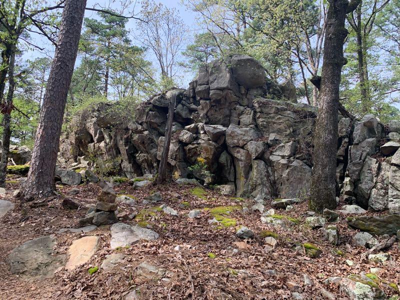 Rugged rock formation surrounded by trees in a wooded area, with patches of moss and fallen leaves on the ground. Iron Mountain mountain bike trail.