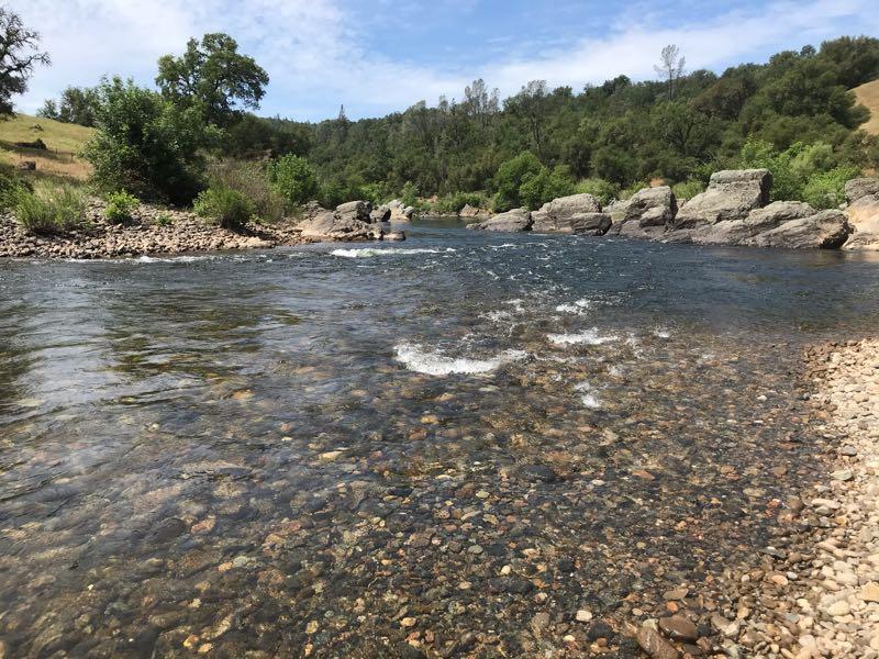 A serene river scene with clear water flowing over smooth stones, surrounded by lush greenery and rocky banks. The sky is partly cloudy, creating a tranquil atmosphere in this natural setting. Cronan Ranch mountain bike trail.