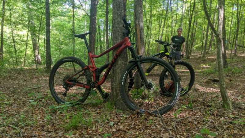 Two mountain bikes are leaning against a tree in a lush green forest. One bike is red and the other is black with yellow accents. In the background, a person stands near the bikes, surrounded by trees and underbrush. The sun is filtering through the leaves, creating a vibrant outdoor atmosphere. Heritage Park mountain bike trail.