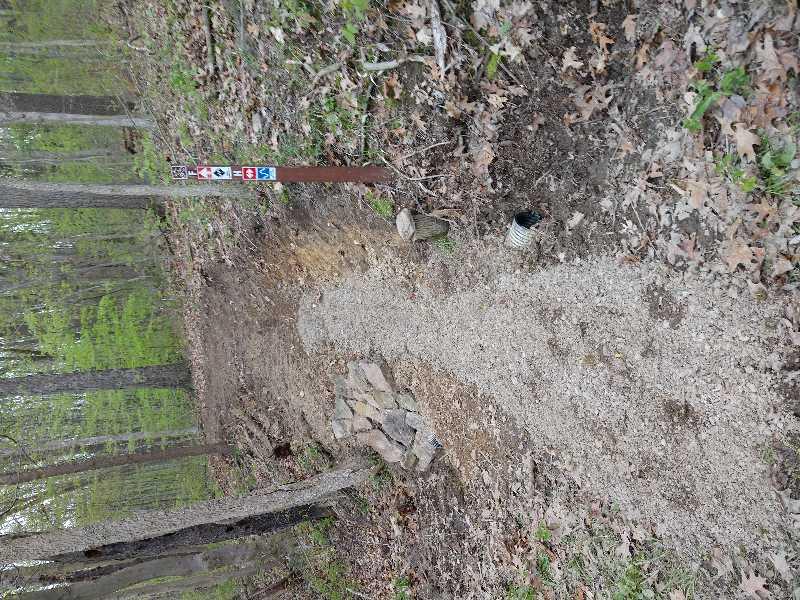 A path leading into a wooded area, marked by a trail sign on the left that indicates directions for hikers. The path is composed of dirt and gravel and is surrounded by lush green foliage and scattered leaves. A small rock formation is visible on the ground along the path. Moraine State Park mountain bike trail.