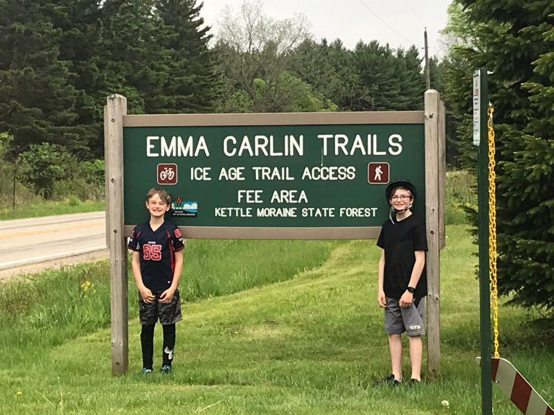 Two children standing in front of a green sign for the Emma Carlin Trails, which includes information on Ice Age Trail access and a note that it is a fee area, located in Kettle Moraine State Forest. Lush greenery surrounds the area, and a road can be seen in the background. Emma Carlin mountain bike trail.