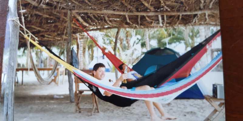 A man relaxing in a colorful hammock under a thatched roof, holding a drink and smiling. In the background, another person sits in a separate hammock, and a tent is visible. The setting appears to be a beach or tropical area with sandy ground and some greenery. Alum Creek Phase II mountain bike trail.