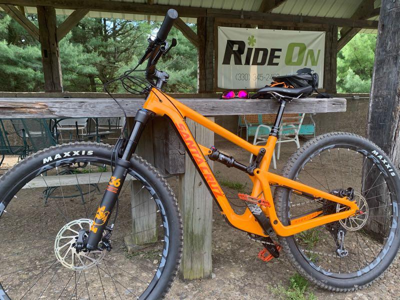 An orange mountain bike is resting against a wooden outdoor table. The bike features thick, knobby tires labeled "Maxxis" and a black front fork with a distinctive design. Nearby, there are sunglasses and a helmet on the table, along with a sign in the background that reads "Ride On." The setting is a forested area with trees visible in the background. Vultures Knob mountain bike trail.