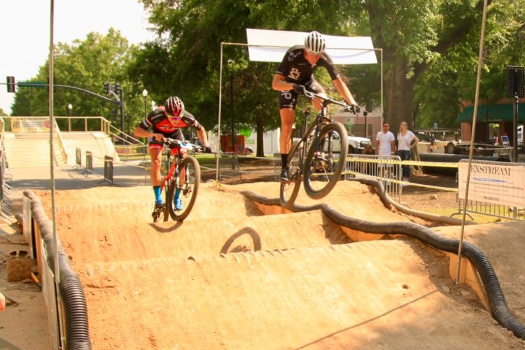 Two mountain bikers are competing on a dirt pump track, maneuvering over a series of bumps. One rider, dressed in red and black, is leaning forward as he pedals, while the other, in black, is airborne above a bump. The background features a park setting with trees and spectators observing the event.