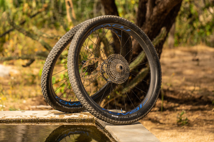 Two mountain bike wheels leaning against a stone edge, showcasing their knobby tires and intricate gears. The background features a blurred natural setting with greenery, and the wheels reflect slightly in a shallow pool of water beside them.