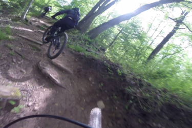 A mountain biker navigating a rocky trail in a lush green forest, with sunlight filtering through the trees in the background. The focus is on the biker as they ride uphill on a dirt path surrounded by vegetation. Richmond Avenue and Forest Hill road mountain bike trail.