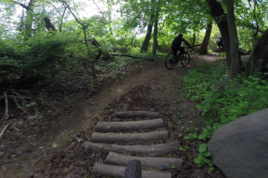 Mountain bikers navigating a wooded trail with a makeshift wooden bridge, surrounded by lush greenery and trees. The perspective shows the trail ahead, emphasizing the natural landscape. Richmond Avenue and Forest Hill road mountain bike trail.