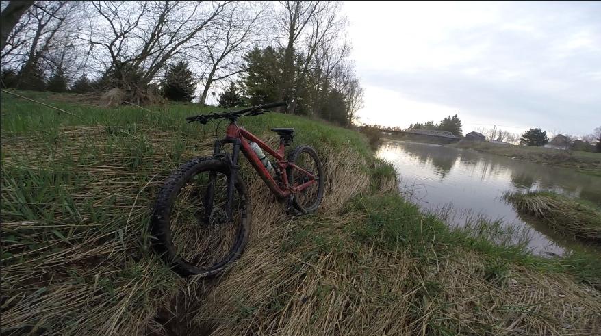 Trek Fuel EX 8 29: A red mountain bike resting on the grassy bank of a calm river, surrounded by trees and an overcast sky.