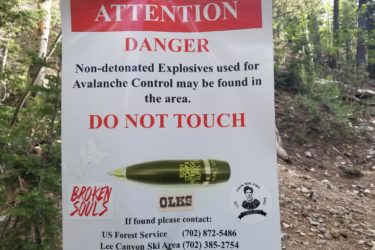 A warning sign in a forested area, stating "Attention Danger" in large red letters. It advises that non-detonated explosives used for avalanche control may be present and instructs not to touch them. The sign features contact numbers for the US Forest Service and Lee Canyon Ski Area for reporting found explosives. Additionally, there are stickers on the sign with phrases like "Broken Souls." The background includes trees and rocky terrain. Bristlecone Trail mountain bike trail.
