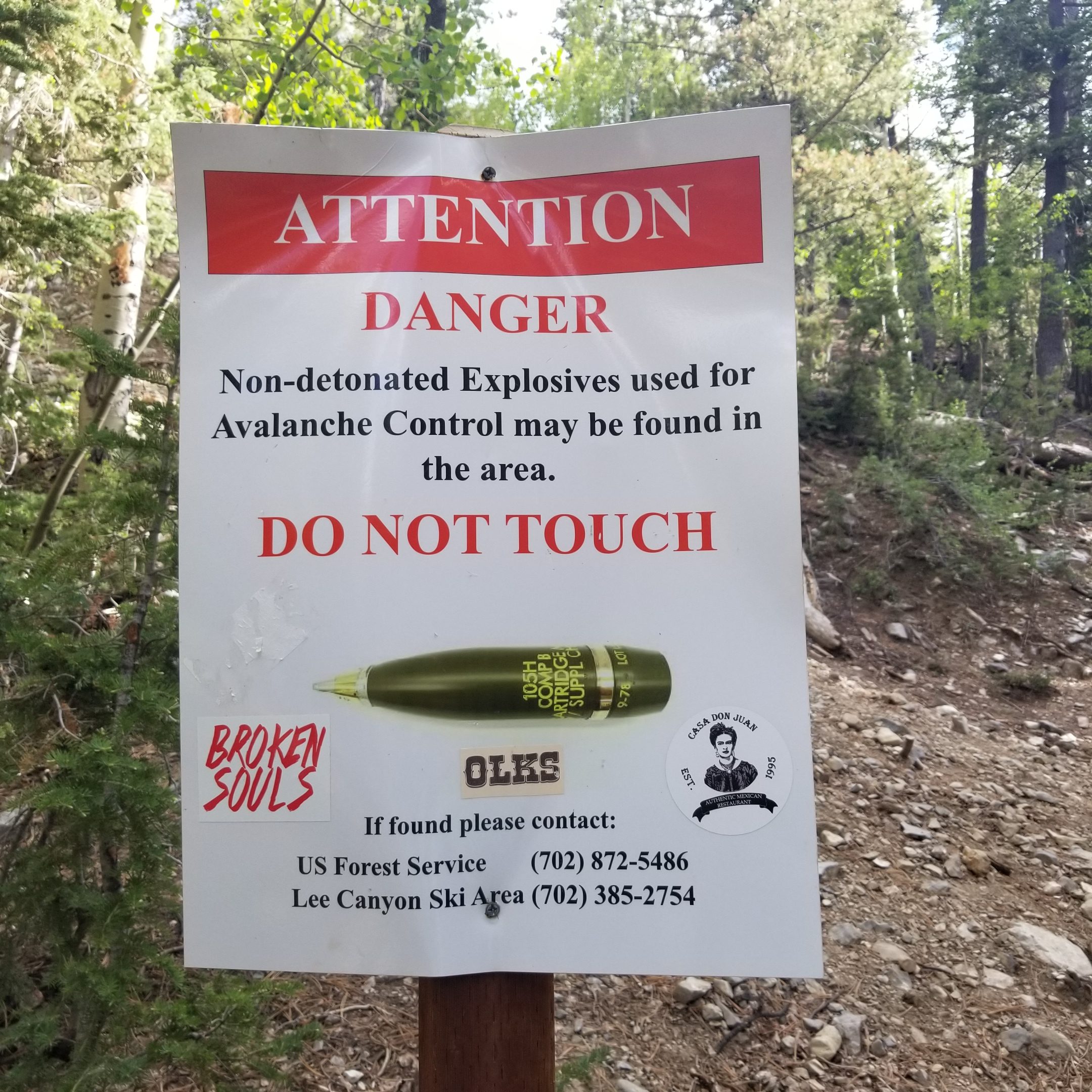 A warning sign in a forested area, stating "Attention Danger" in large red letters. It advises that non-detonated explosives used for avalanche control may be present and instructs not to touch them. The sign features contact numbers for the US Forest Service and Lee Canyon Ski Area for reporting found explosives. Additionally, there are stickers on the sign with phrases like "Broken Souls." The background includes trees and rocky terrain. Bristlecone Trail mountain bike trail.